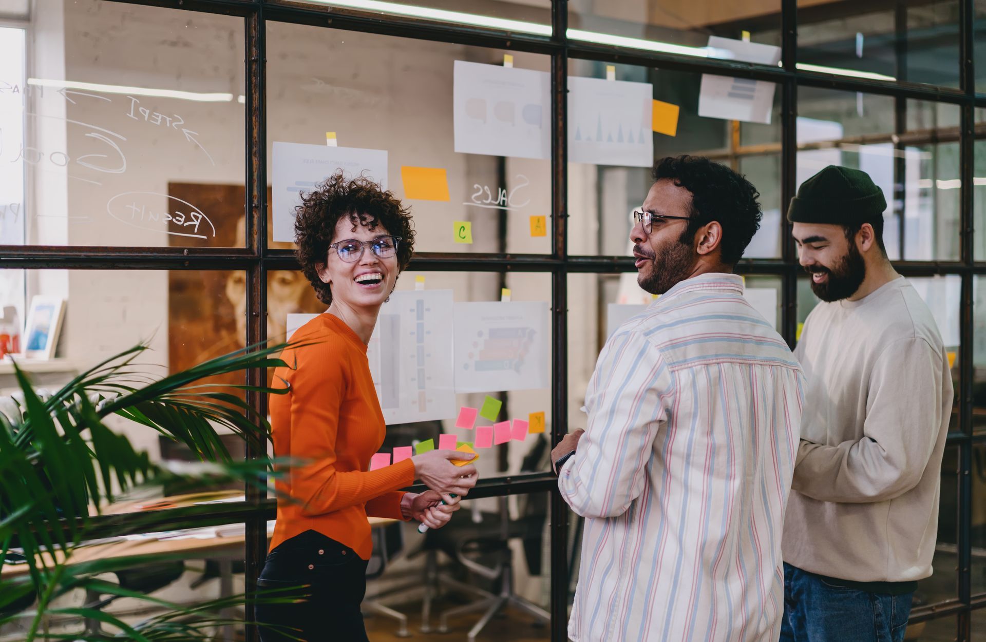 Three people in an office, laughing and discussing ideas near a glass wall with sticky notes.