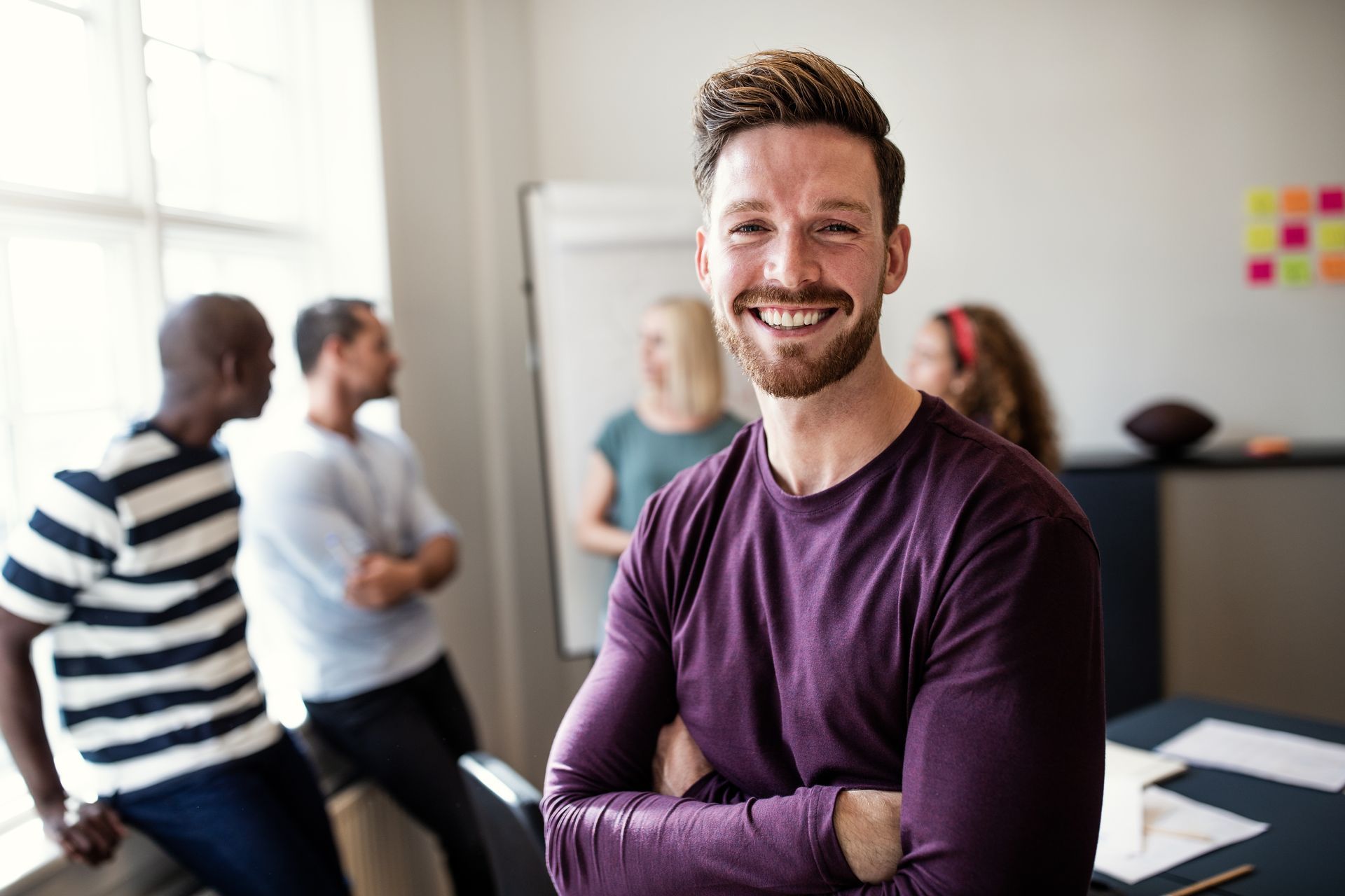 Man with arms crossed smiling, others blurred in background.