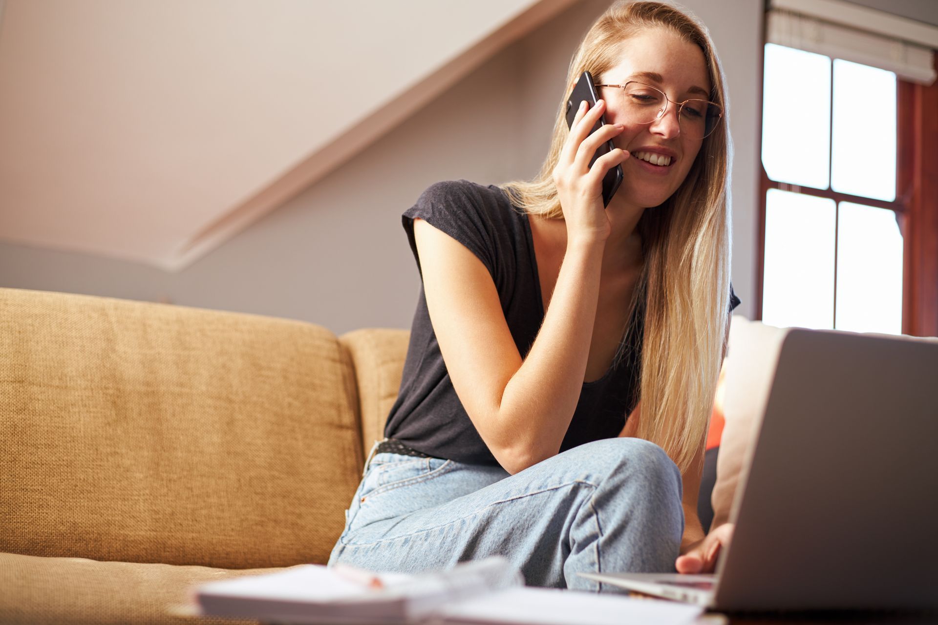 Woman wearing glasses smiles while talking on phone and using a laptop on a sofa.