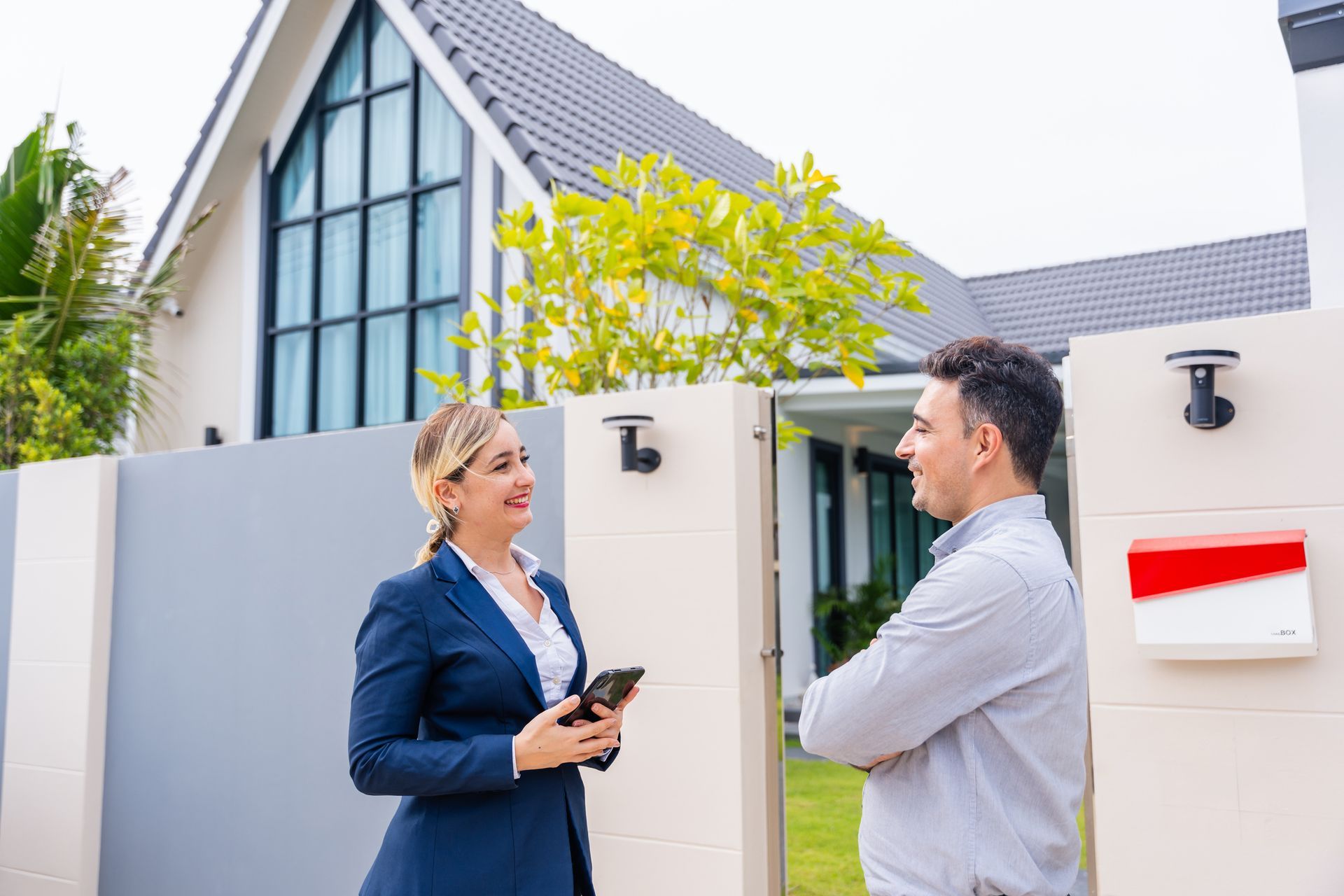 Real estate agent in a blue suit holding a phone conversing with a man at the entrance of a modern house.