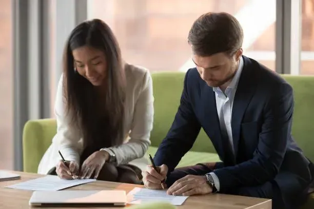 A woman and a man in business attire sign documents at a table, likely during a meeting.