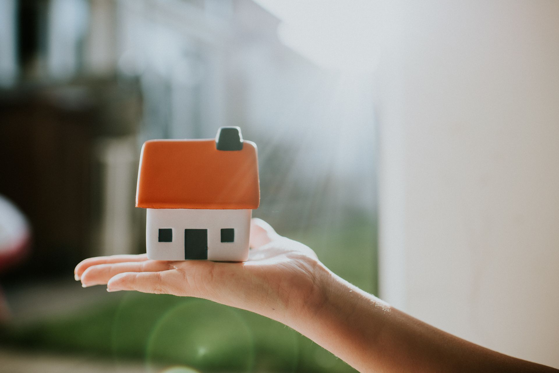 Hand holding a small white house with an orange roof; sunlight shines through a window in the background.