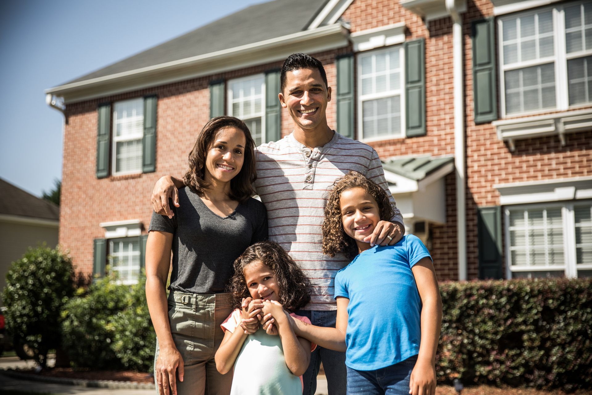 Family of four smiling in front of a brick house on a sunny day.