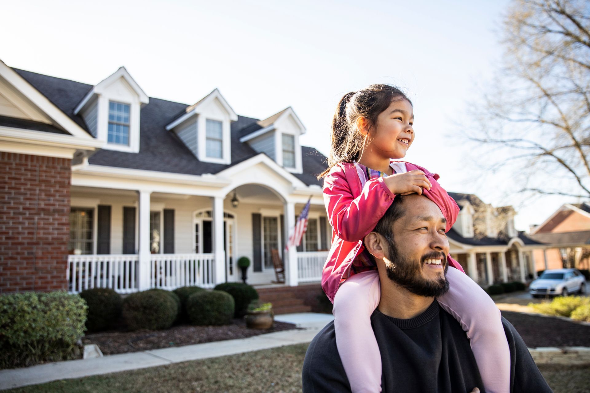 A man carries a young girl on his shoulders in front of a two-story house. The girl smiles and looks ahead.