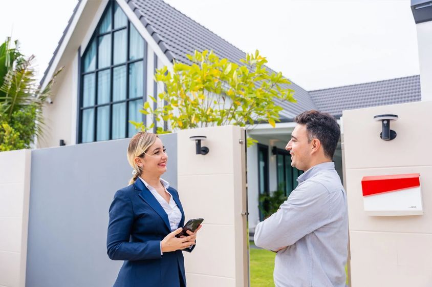 Real estate agent in a blue suit holding a phone conversing with a man at the entrance of a modern house.