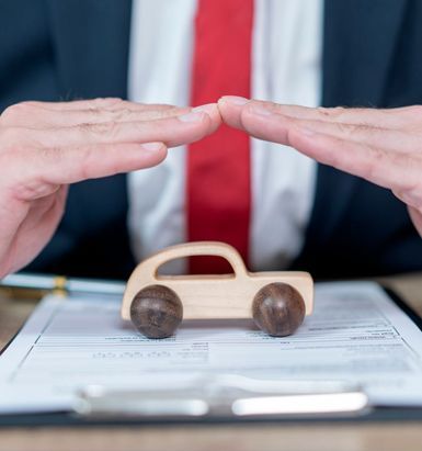 Person in a suit protects a small wooden car with their hands, placed over a contract on a desk.