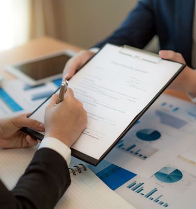Two men in suits at a desk; one signing a document held by the other, surrounded by financial graphs.