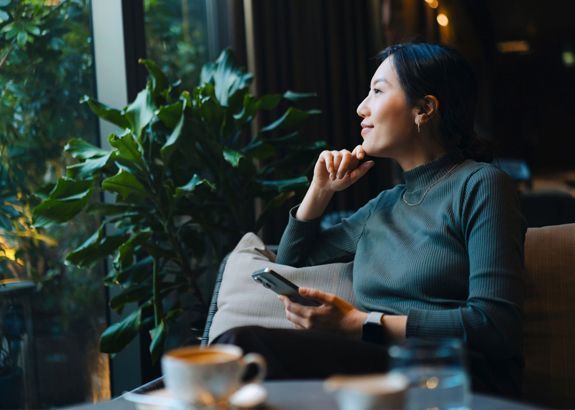 Woman looking out a window, holding a phone and resting her chin on her hand. She sits indoors near a large green plant.