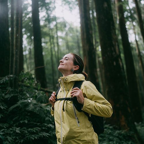 A woman in a yellow jacket is standing in the woods with her eyes closed.