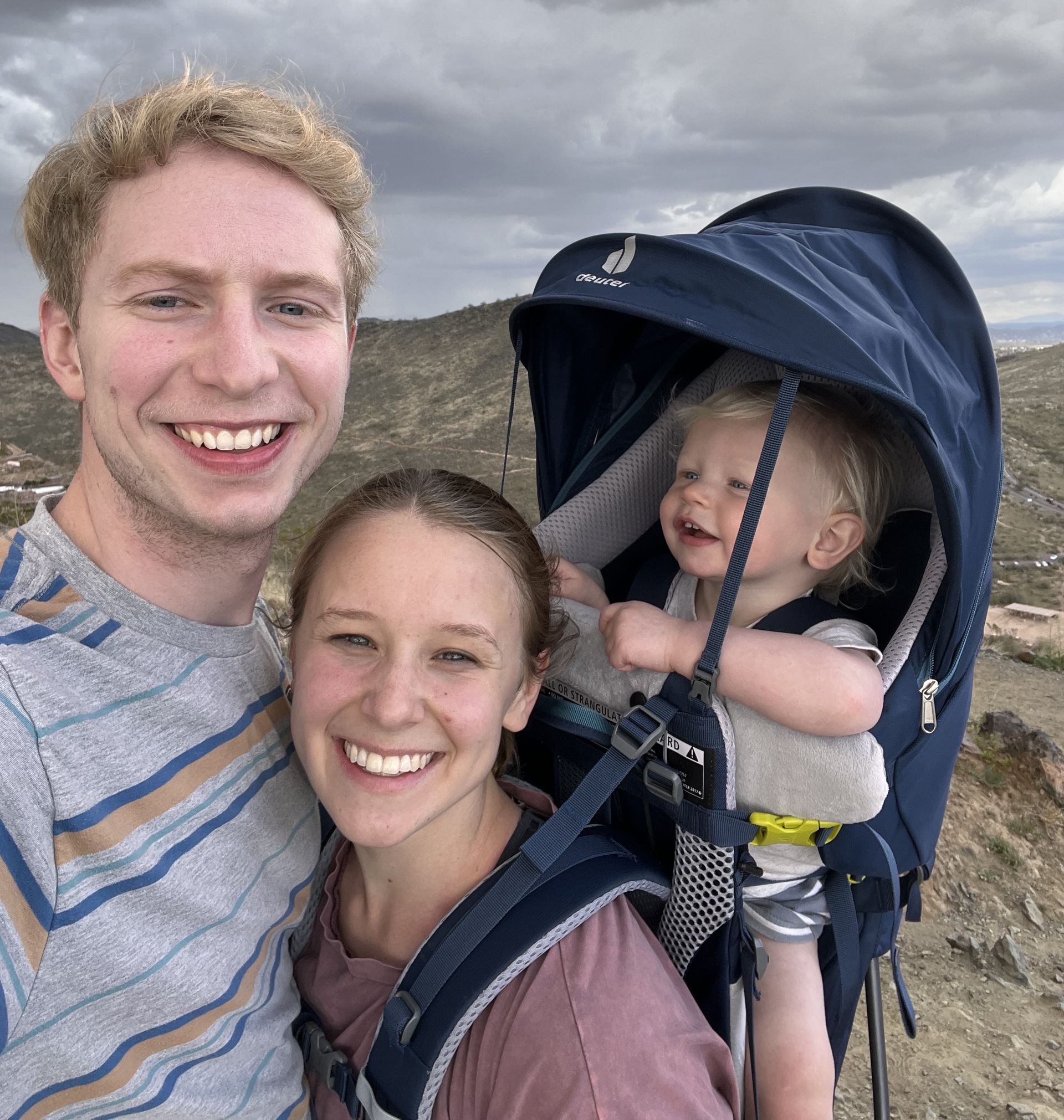 Smiling family selfie outdoors, with a baby in a navy backpack carrier under cloudy skies.
