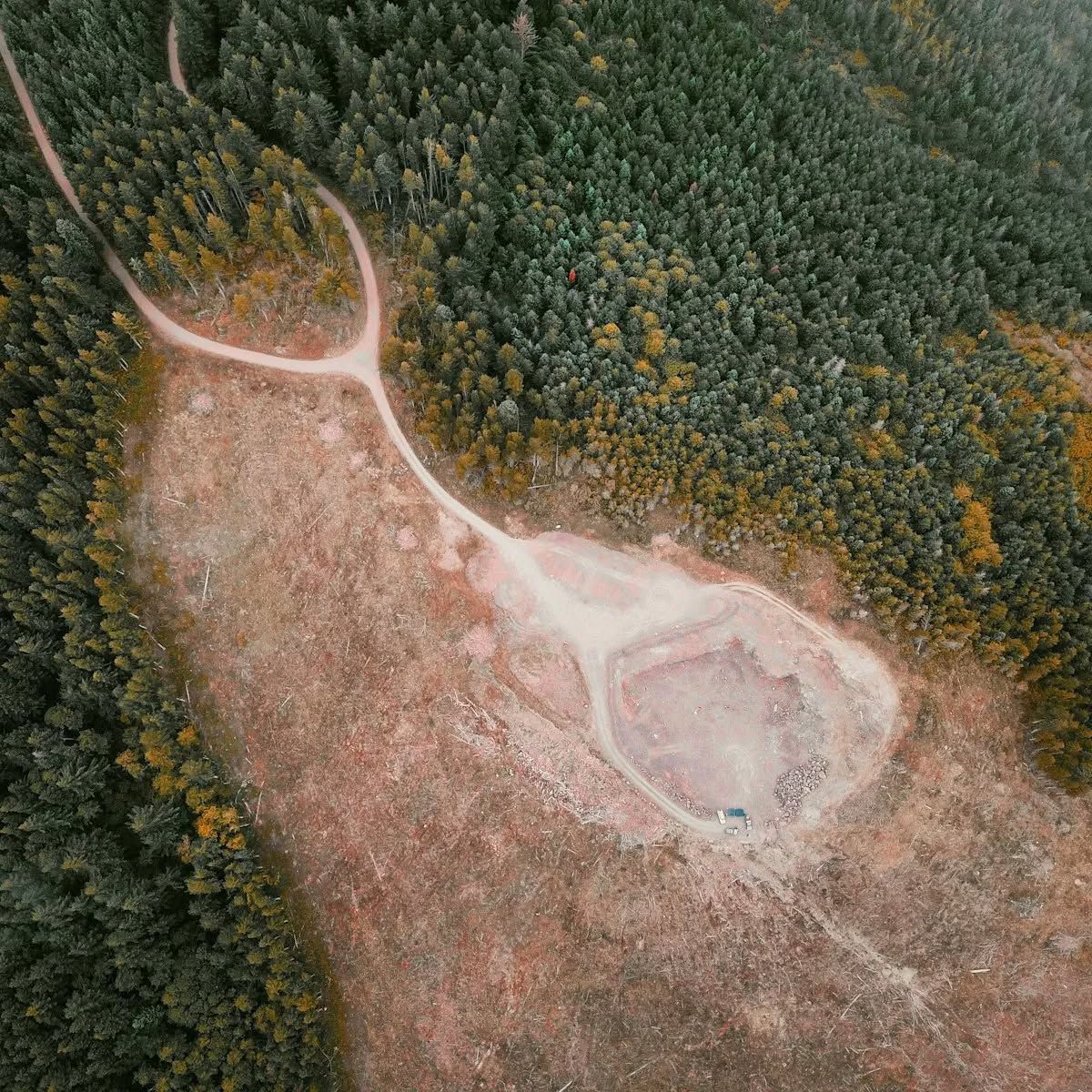 An aerial view shows a Y-shaped dirt road leading into a cleared, circular quarry area surrounded by a dense green forest.