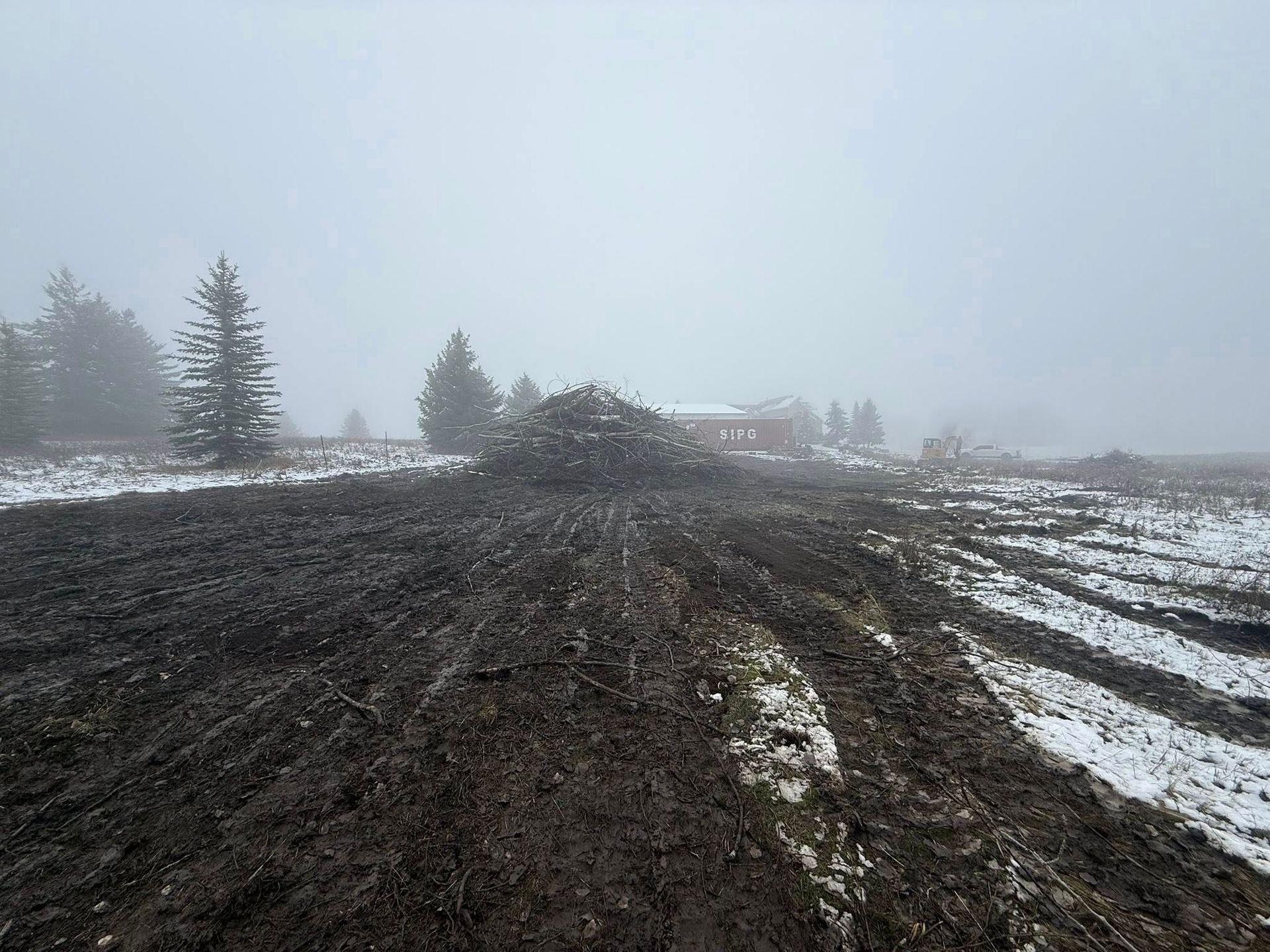 A muddy, plowed field with patches of snow under a foggy sky, featuring a large mound of dark soil in the distance.