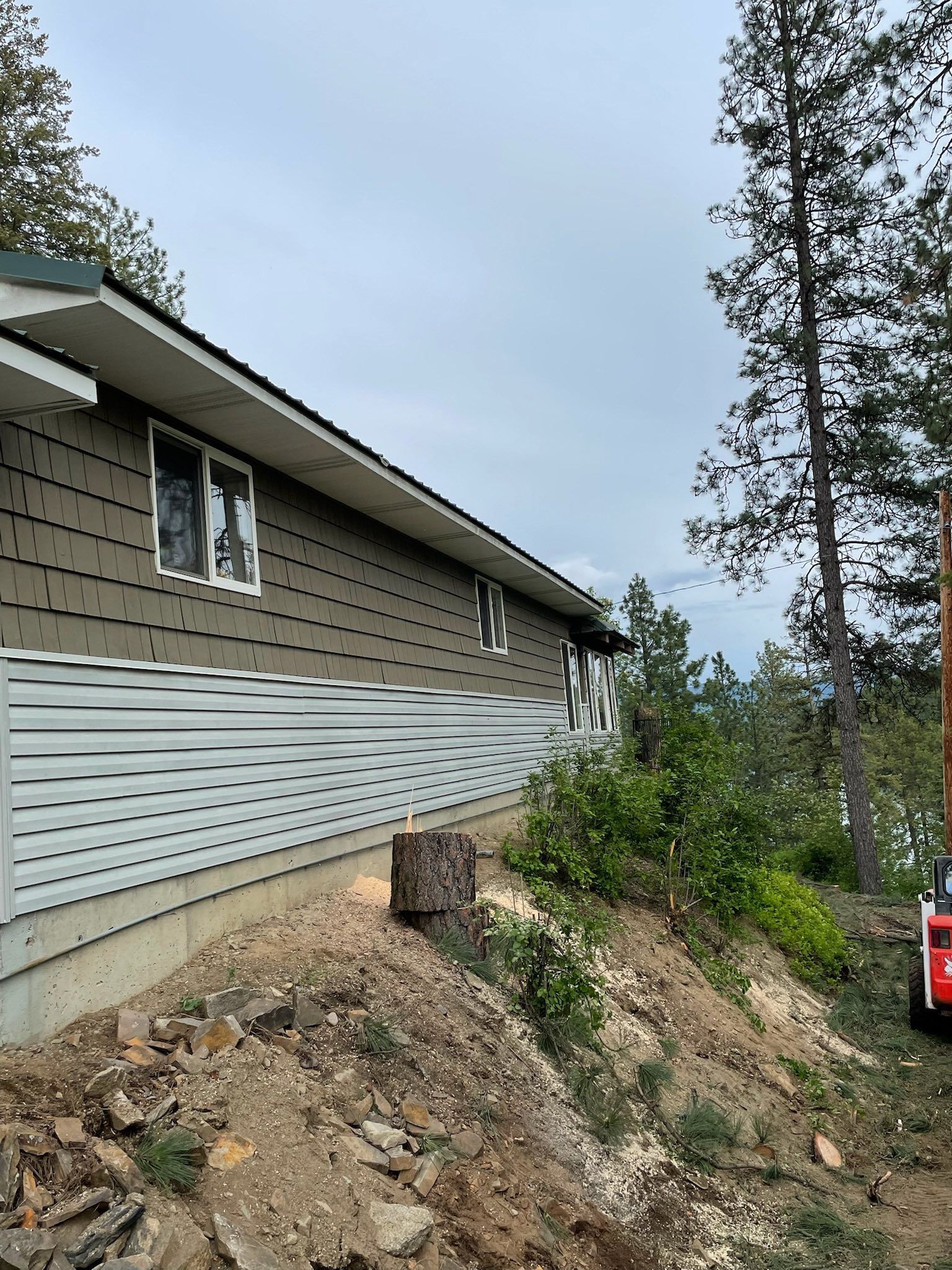 A brown house with white horizontal siding on a slope next to trees, with a piece of construction equipment at the edge.