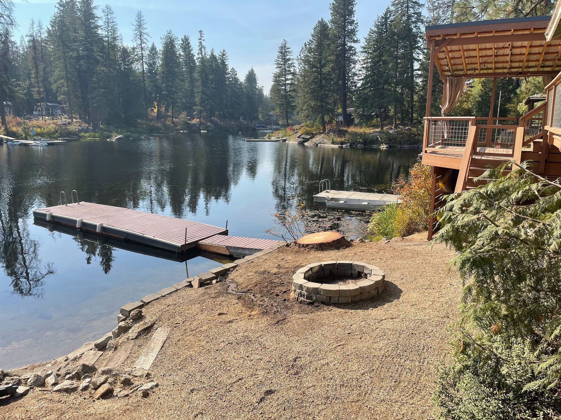 A calm lakefront scene with a floating dock, a stone fire pit, and a wooden deck under tall pine trees.
