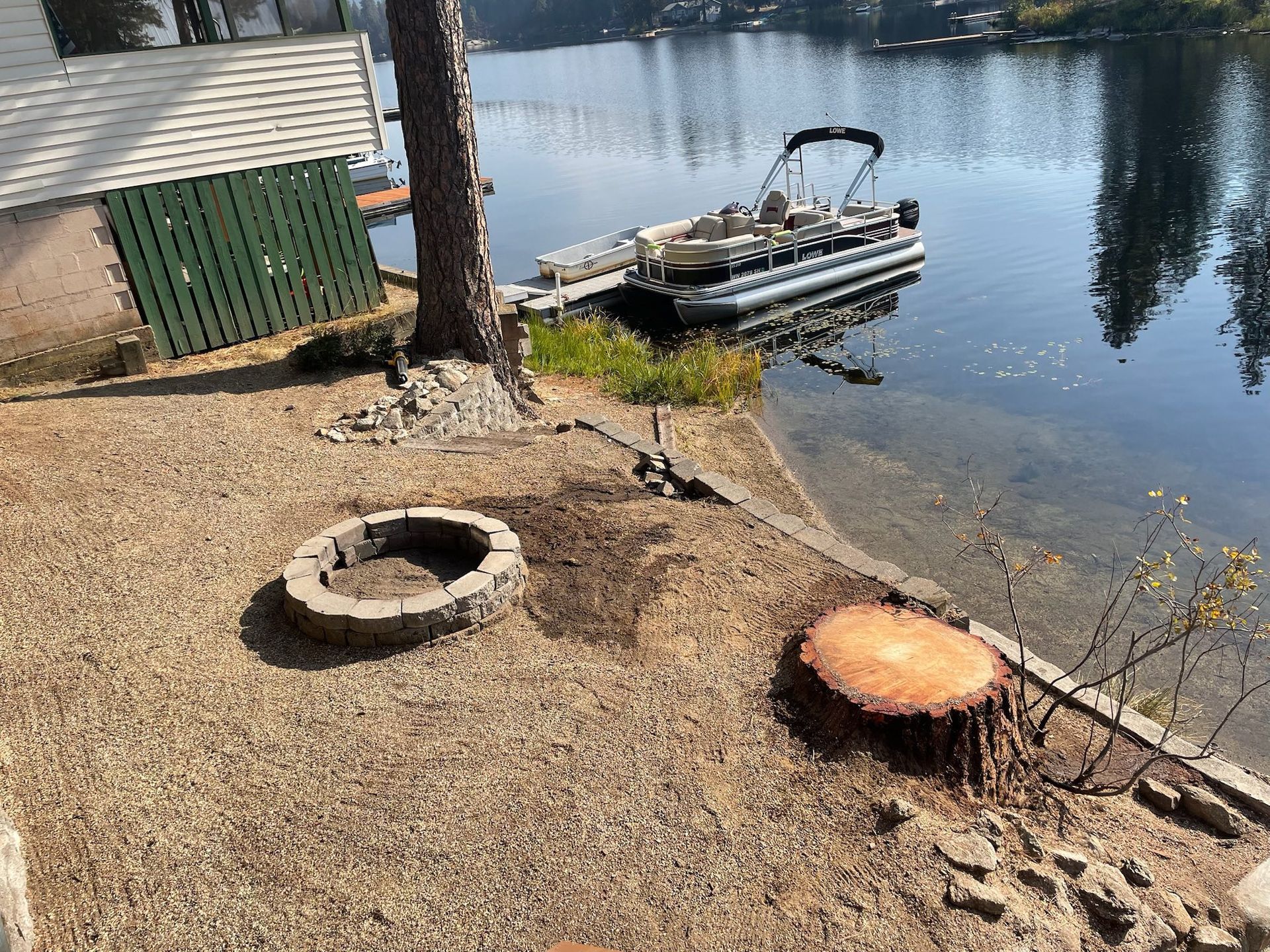 A lakefront backyard with a stone fire pit, a large tree stump, and a pontoon boat docked in the water.