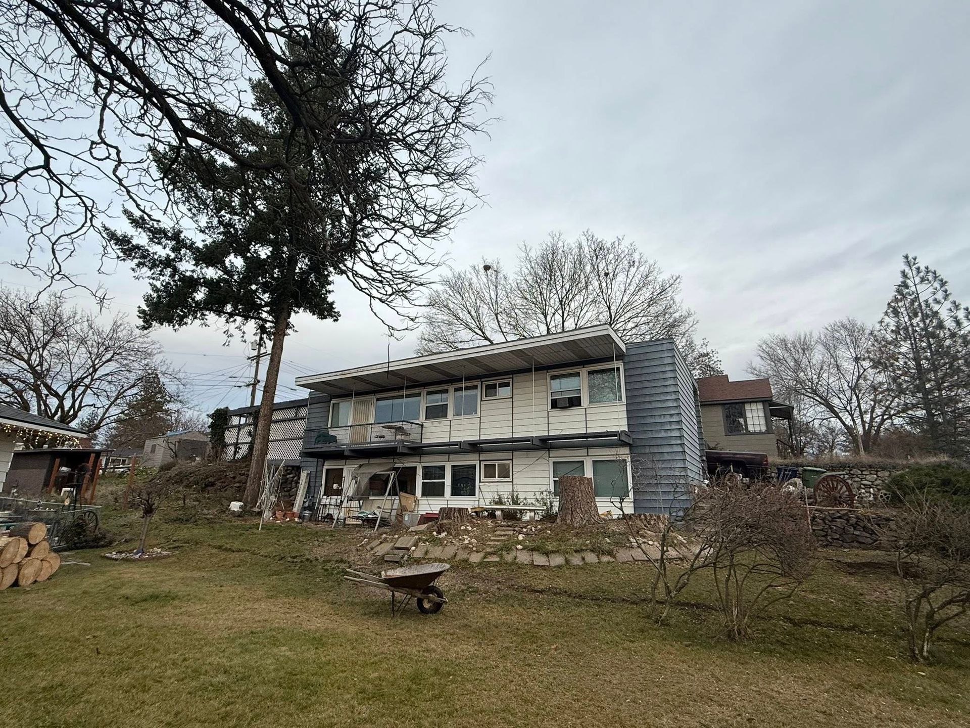 A two-story house under renovation, featuring exposed framing, unfinished siding, and a large tree in a grassy yard.
