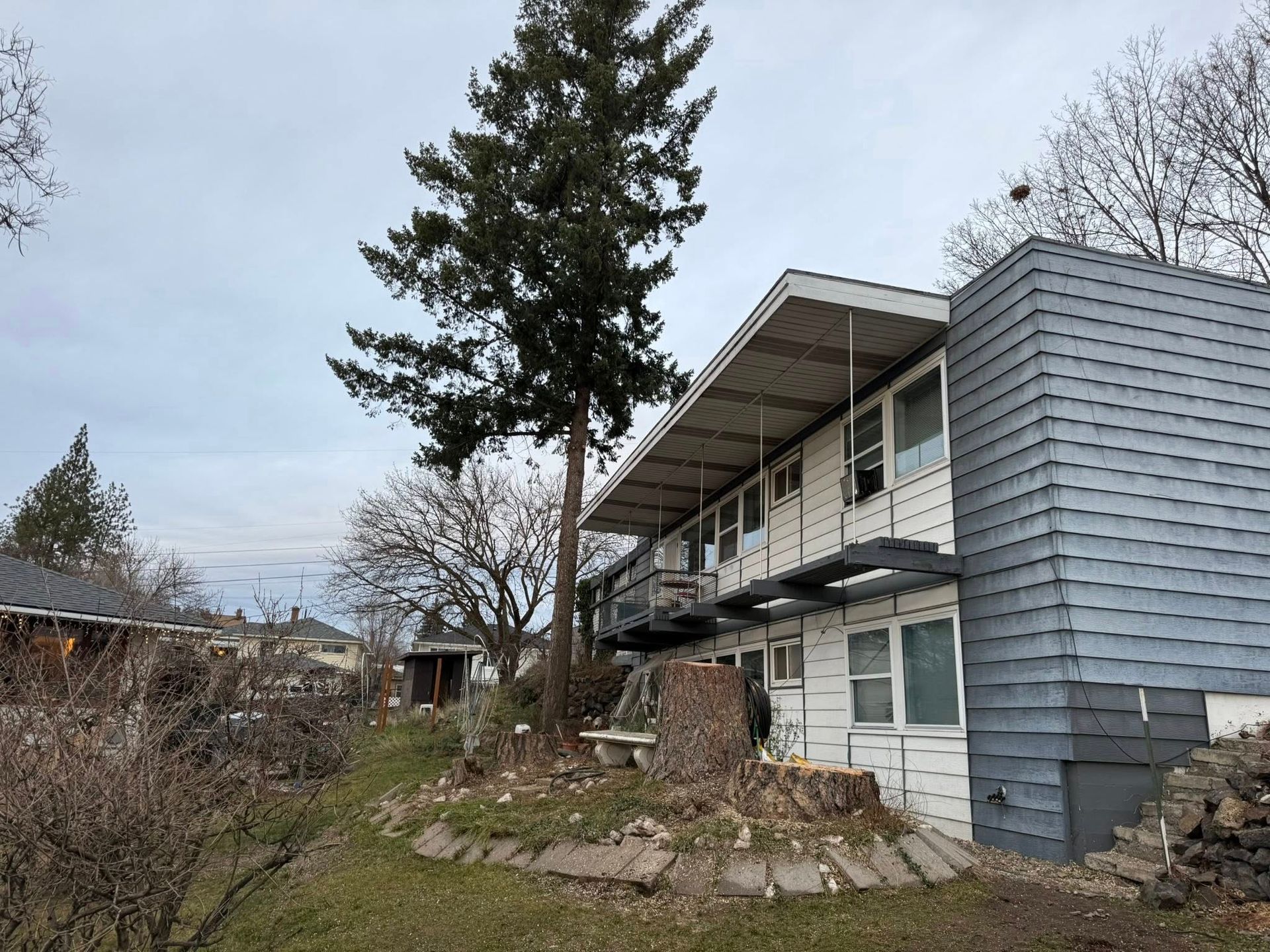 Two-story apartment building with a flat roof, white siding, and a prominent blue-grey textured end wall near a pine tree.