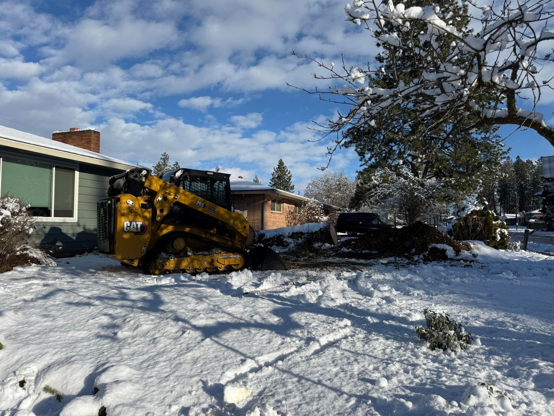A yellow Caterpillar compact track loader sits in a snow-covered yard near a house, working on dirt and debris removal.