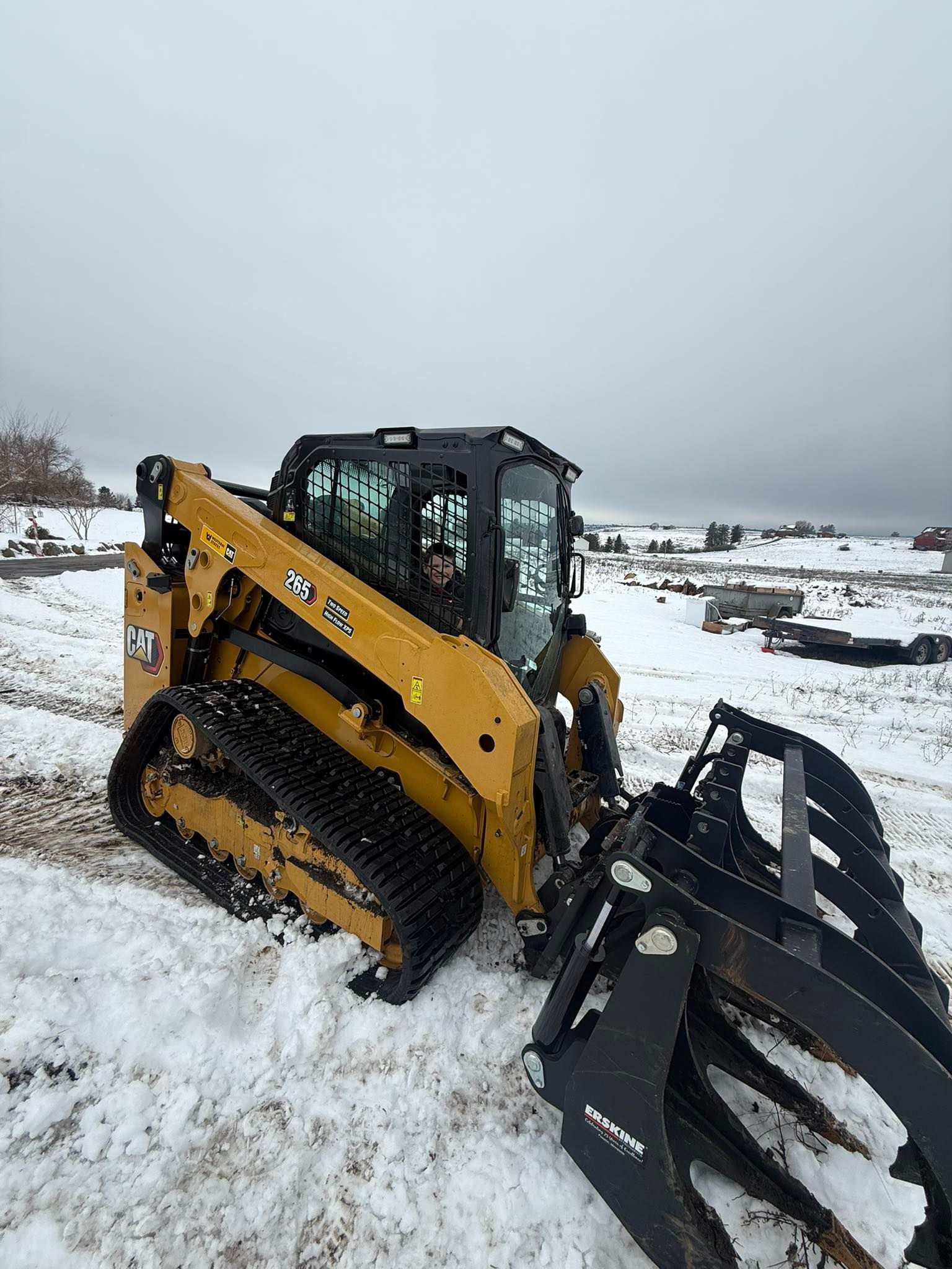 A yellow Caterpillar compact track loader with a black grapple attachment parked in a snowy, rural field.