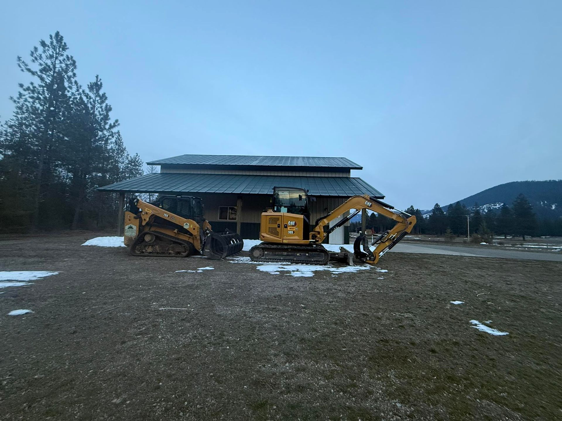 Two yellow Caterpillar construction machines parked on a dirt lot in front of a building with a snowy roof.