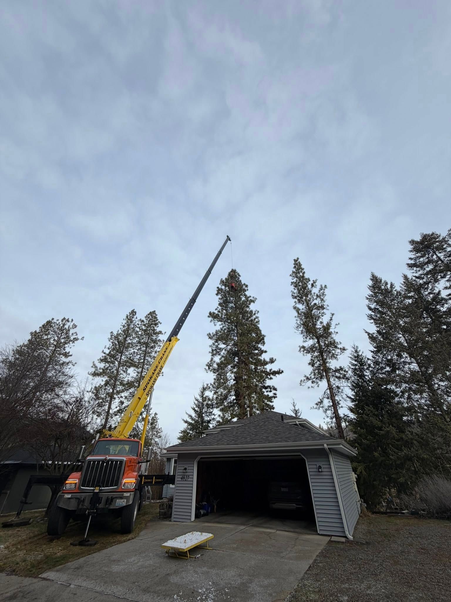 A yellow crane is positioned next to a gray garage, extending its boom upward toward tall pine trees.