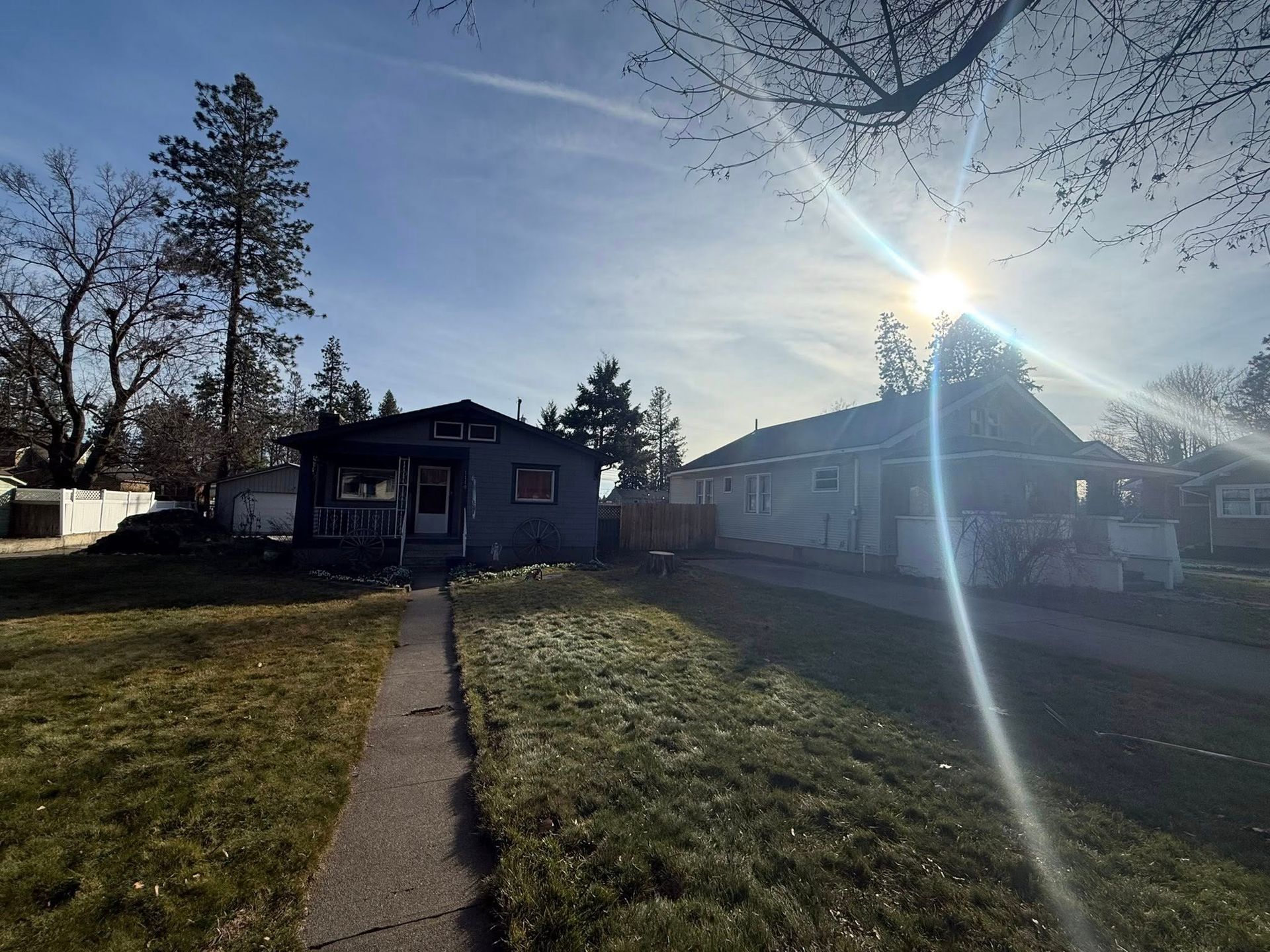 Two houses sit under a bright, lens-flared sun on a clear day with dry, grassy lawns and bare trees in the background.