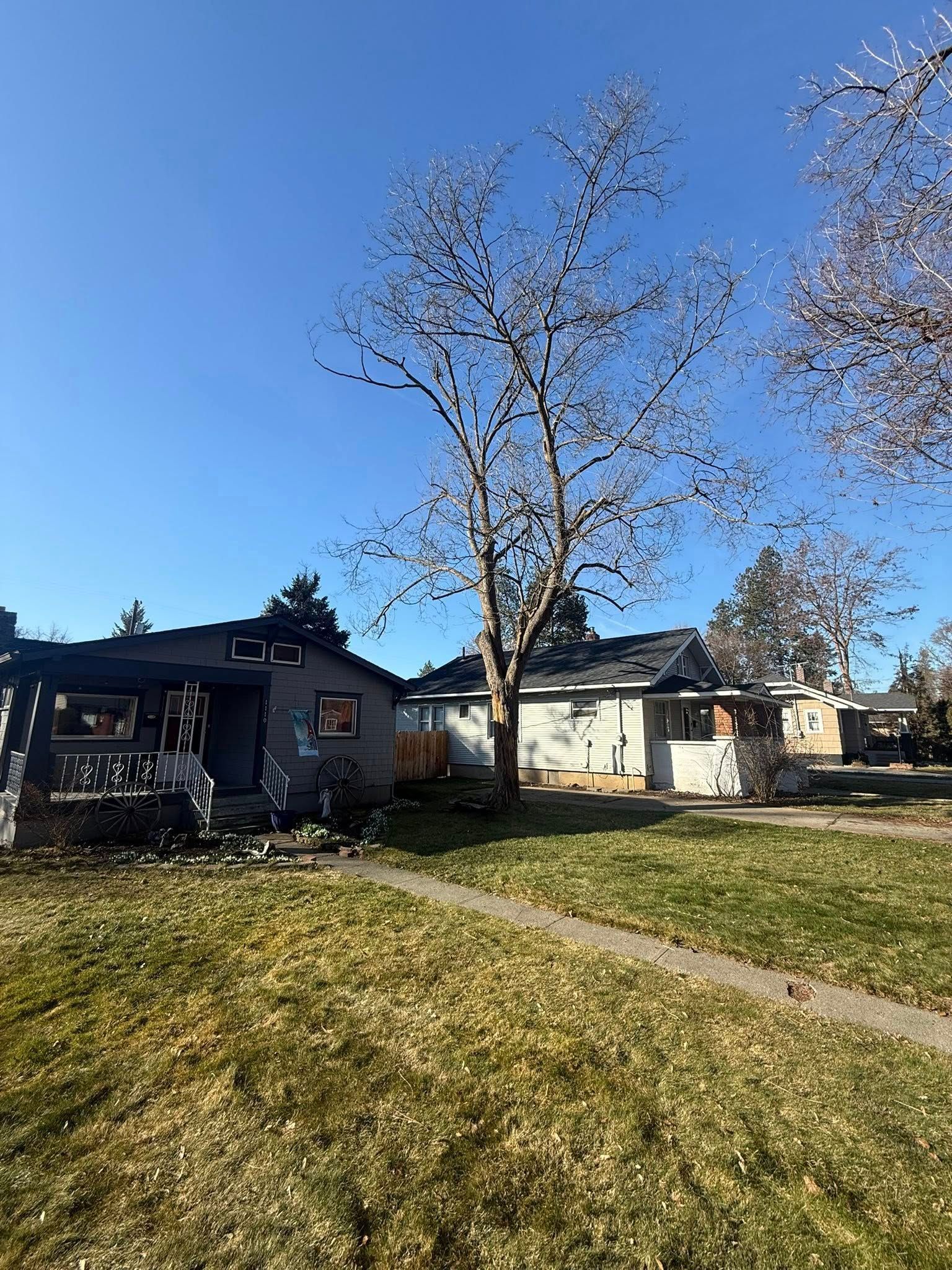 A partially burned house with charred siding stands next to a neighboring home under a bright blue sky.