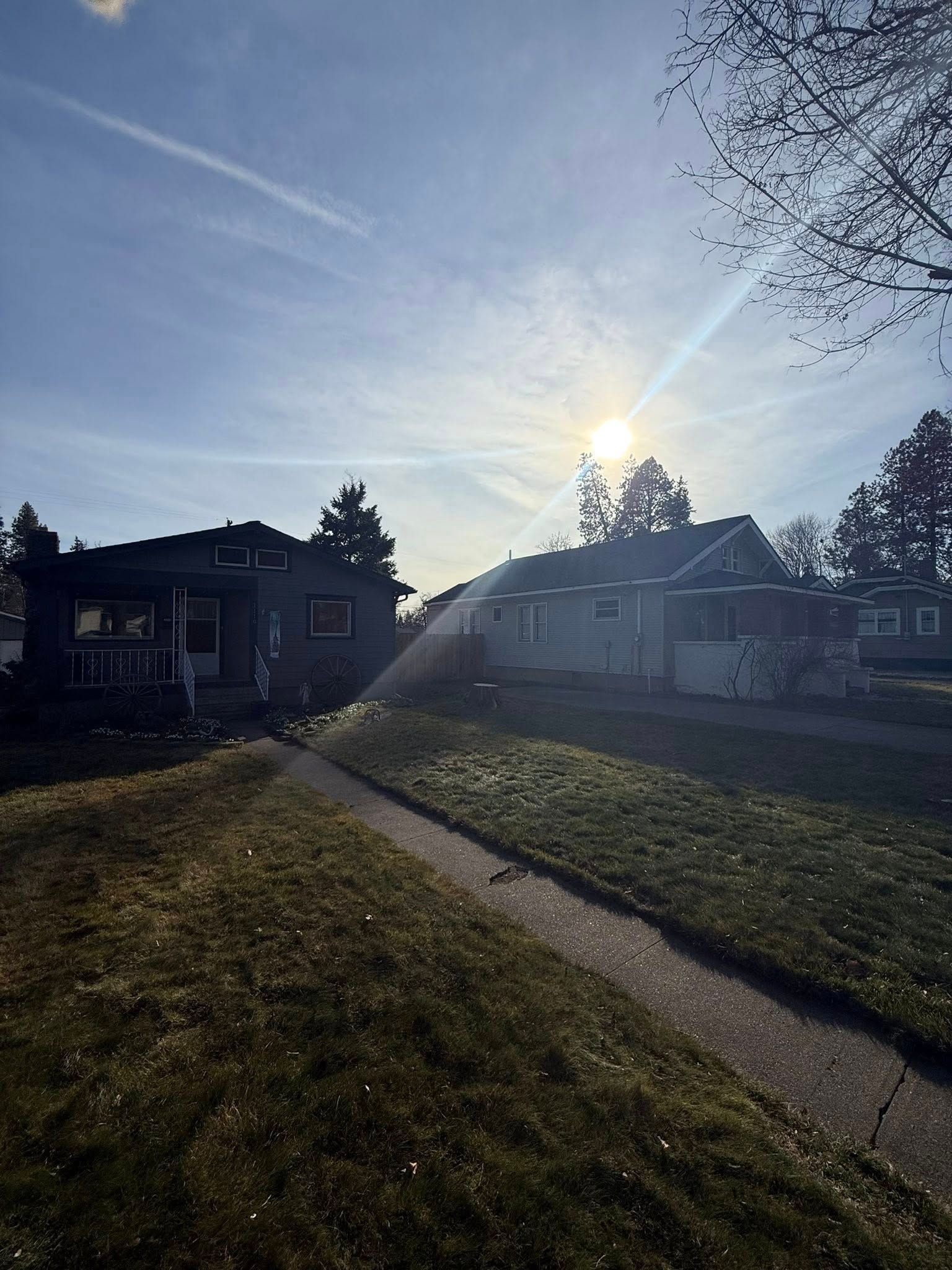 A sunlit neighborhood scene with two houses, a sidewalk, and patchy grass under a clear blue sky.