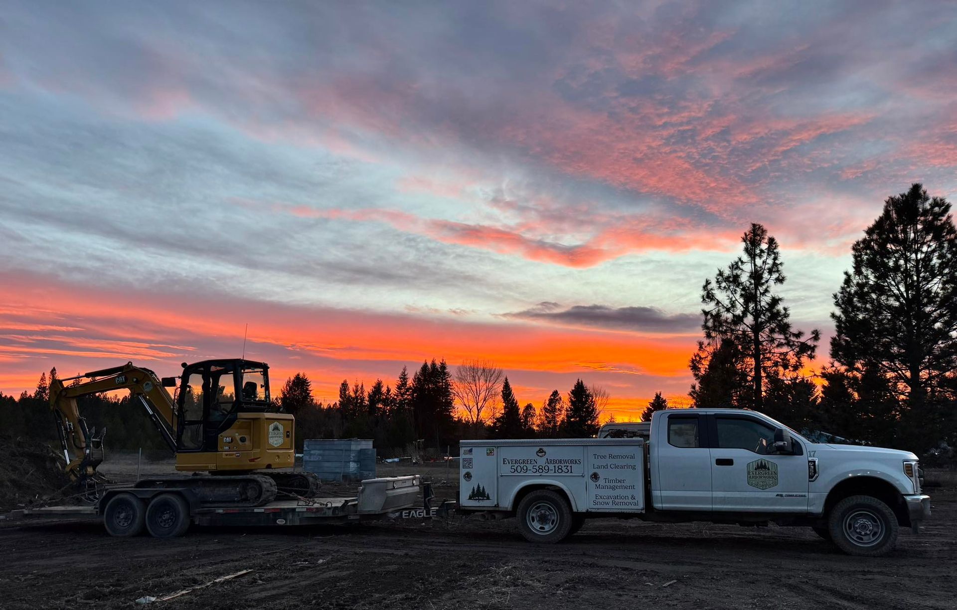 A white work truck tows a yellow excavator on a flatbed trailer against a vibrant orange and purple sunset.