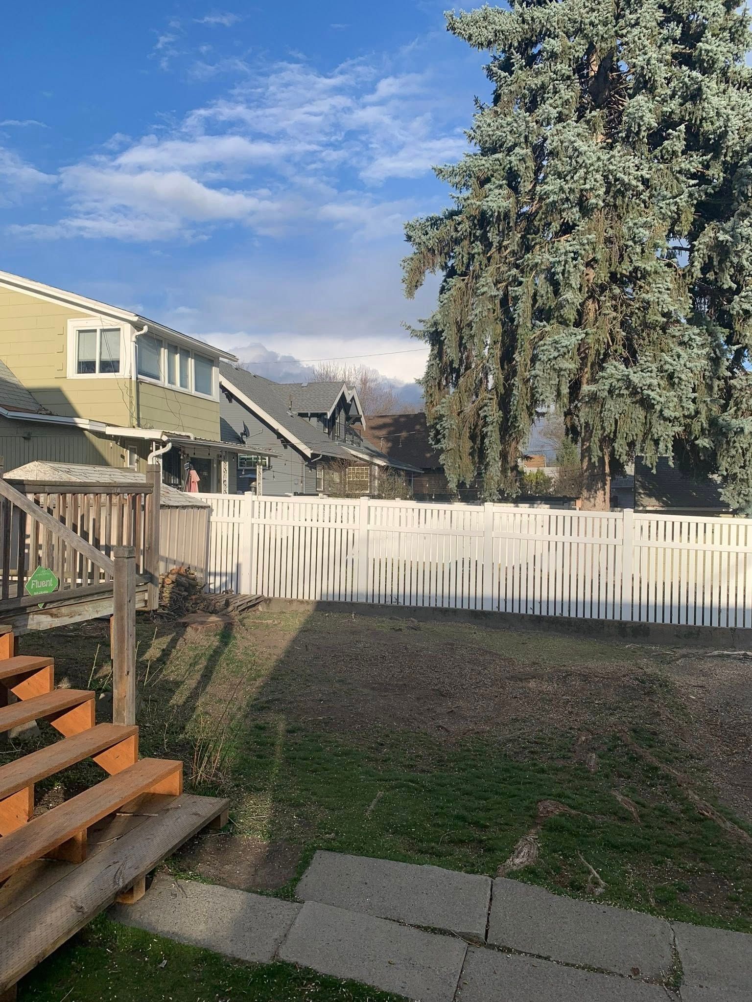 A backyard with wooden stairs in the foreground leading to a lawn, a white picket fence, and houses under a blue sky.