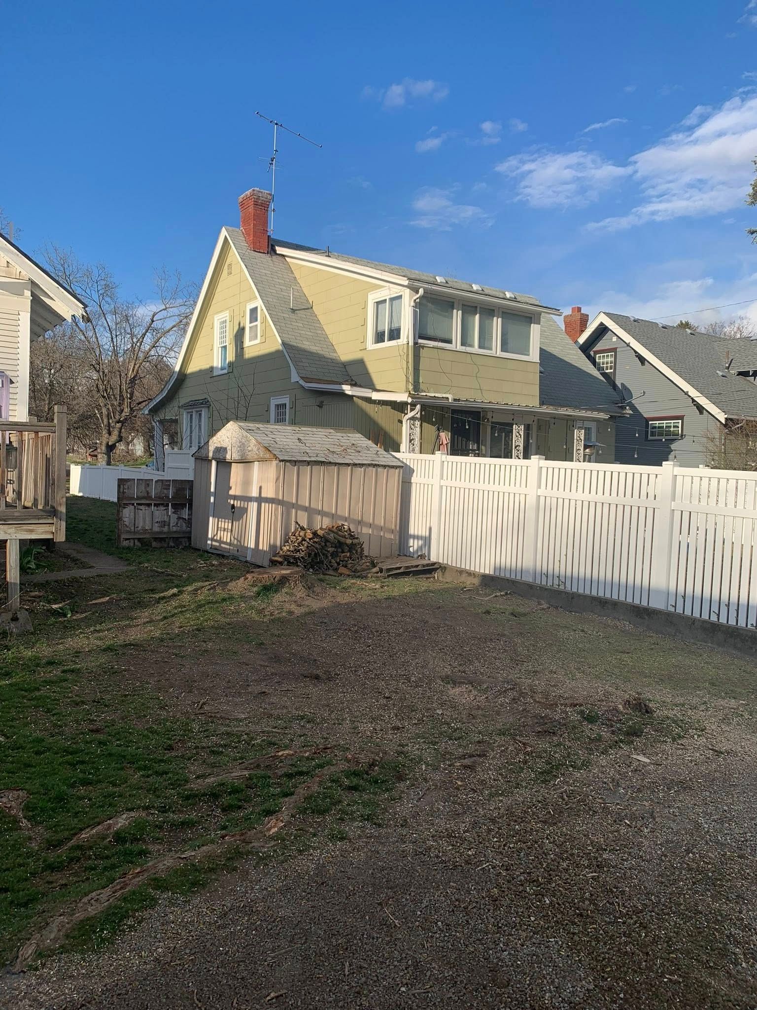 A yellow two-story house with a red chimney sits behind a white picket fence, viewed from a gravel driveway on a sunny day.
