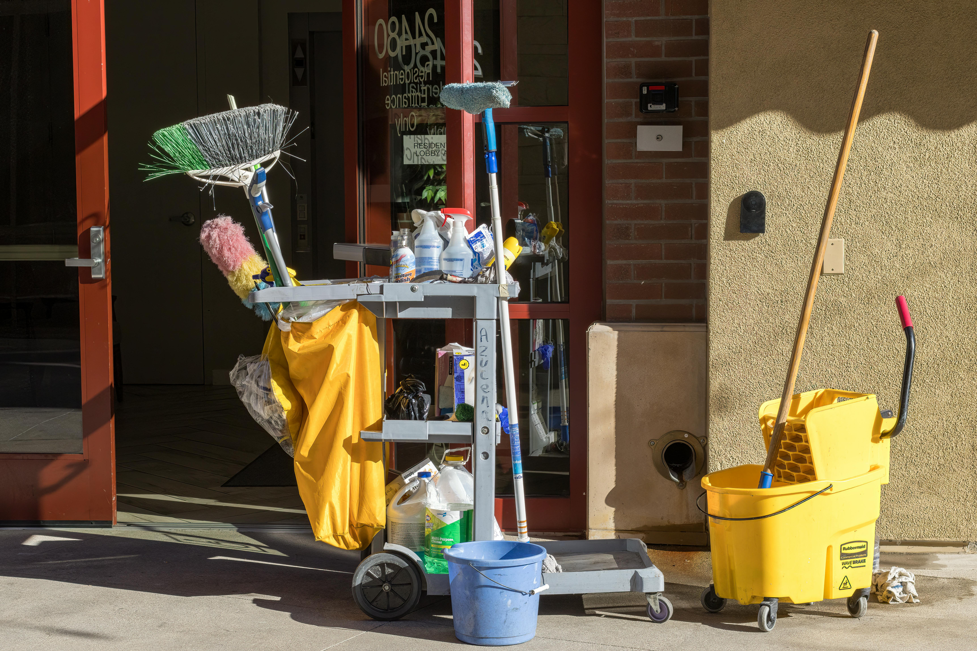 Cleaning cart with supplies next to a building entrance. Yellow bucket, mops, brooms, and cleaning solutions.