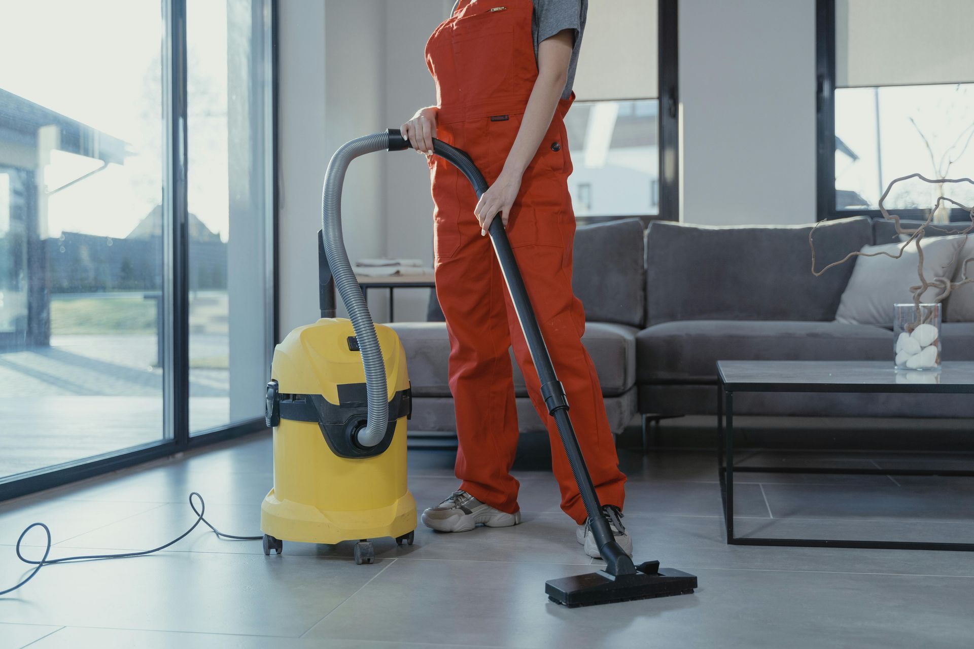 Person in red overalls vacuums a room with large windows, a yellow vacuum cleaner, and gray furniture.