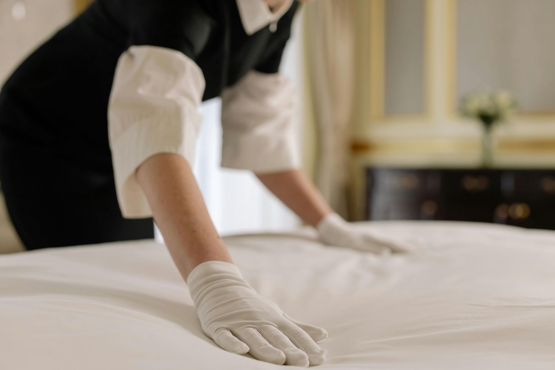 Maid in uniform making a bed, wearing gloves, in a hotel room.