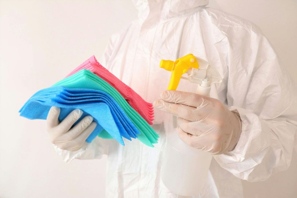 Person in protective suit holding cleaning supplies: spray bottle and colored cloths.