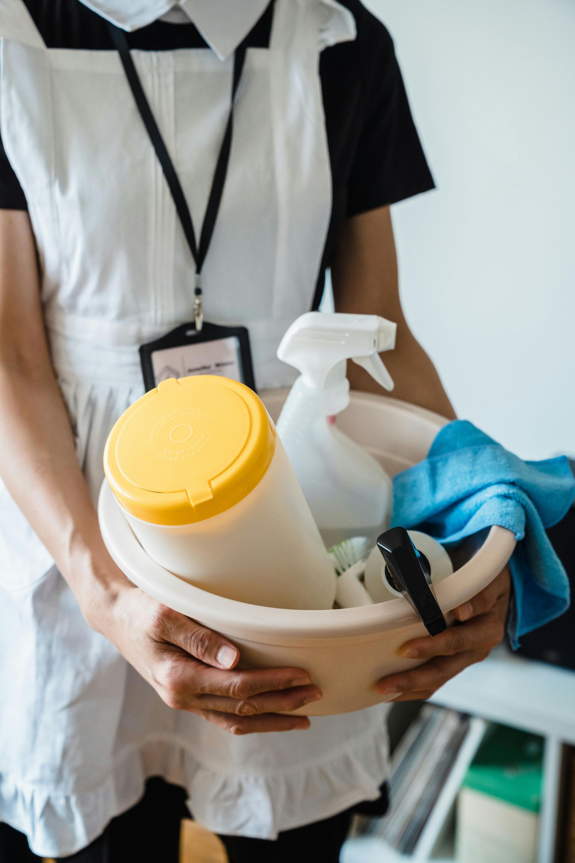 Person in a maid outfit holds a cleaning bucket with supplies: wipes, spray bottles, and a rag.