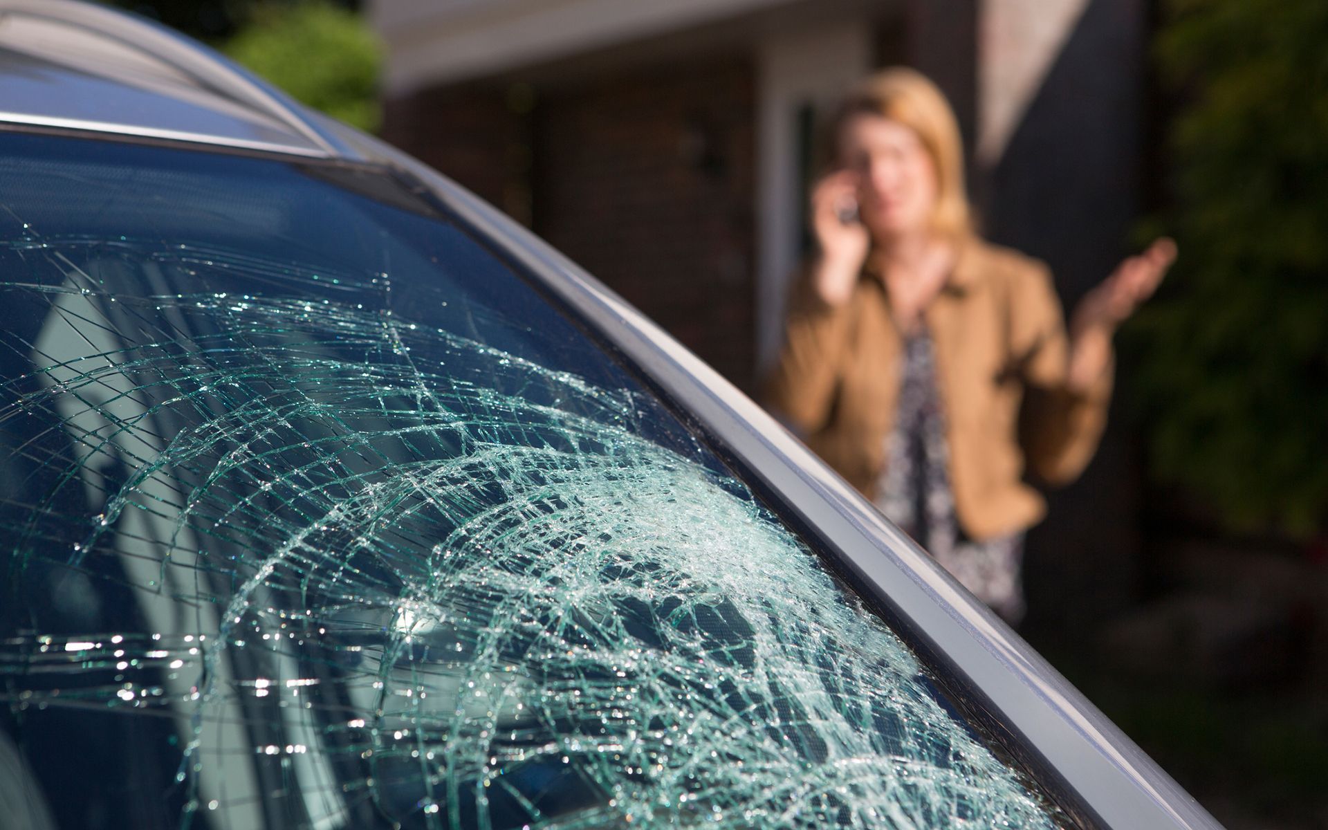 A woman is talking on a cell phone in front of a car with a broken windshield.