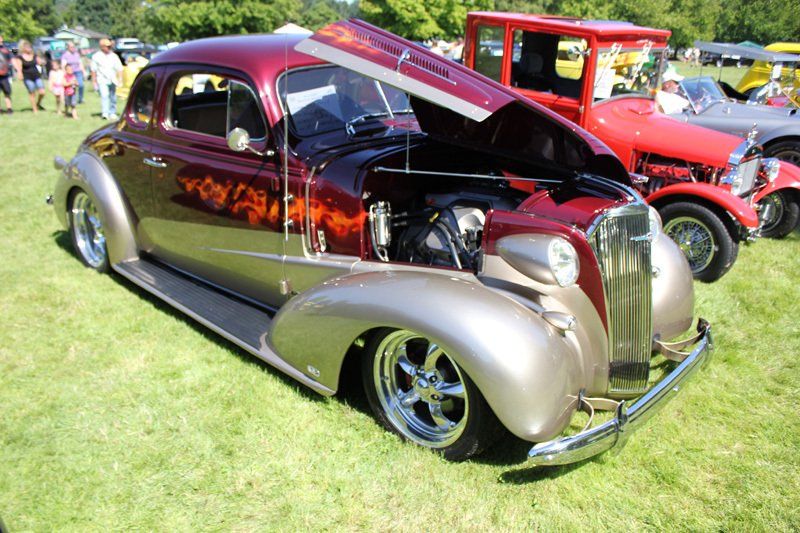A red car with the hood up is parked in the grass at a car show.