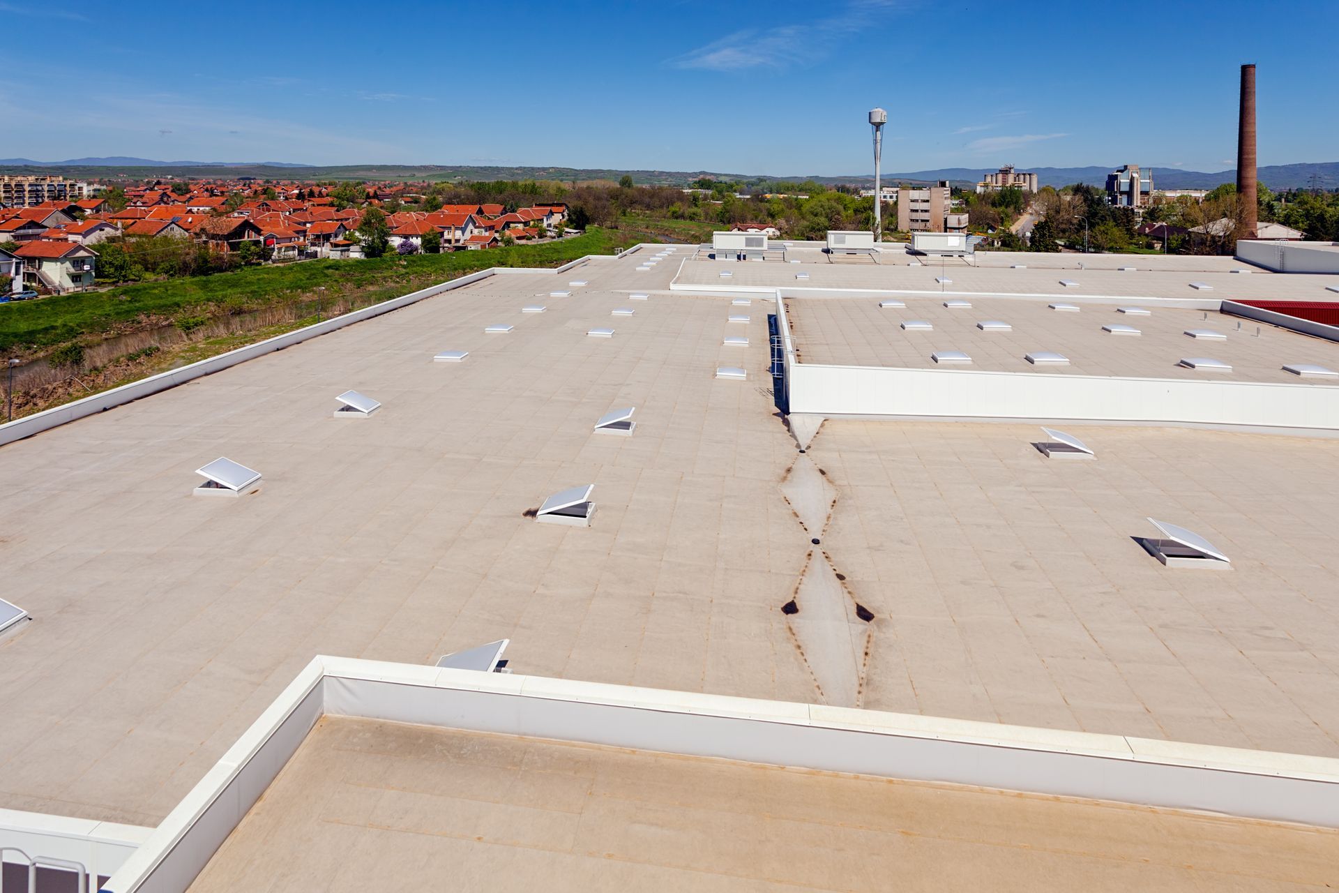 Aerial view of a large flat industrial roof with skylights and surrounding buildings under a clear blue sky.