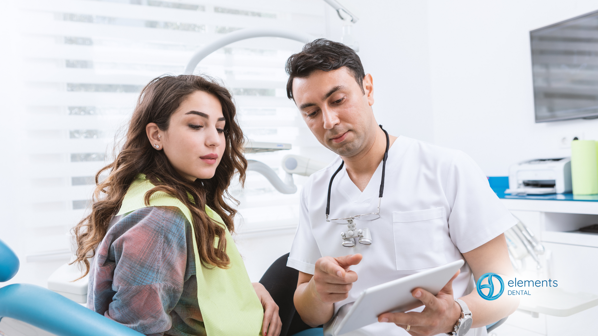 A dentist is talking to a patient in a dental chair while looking at a tablet.