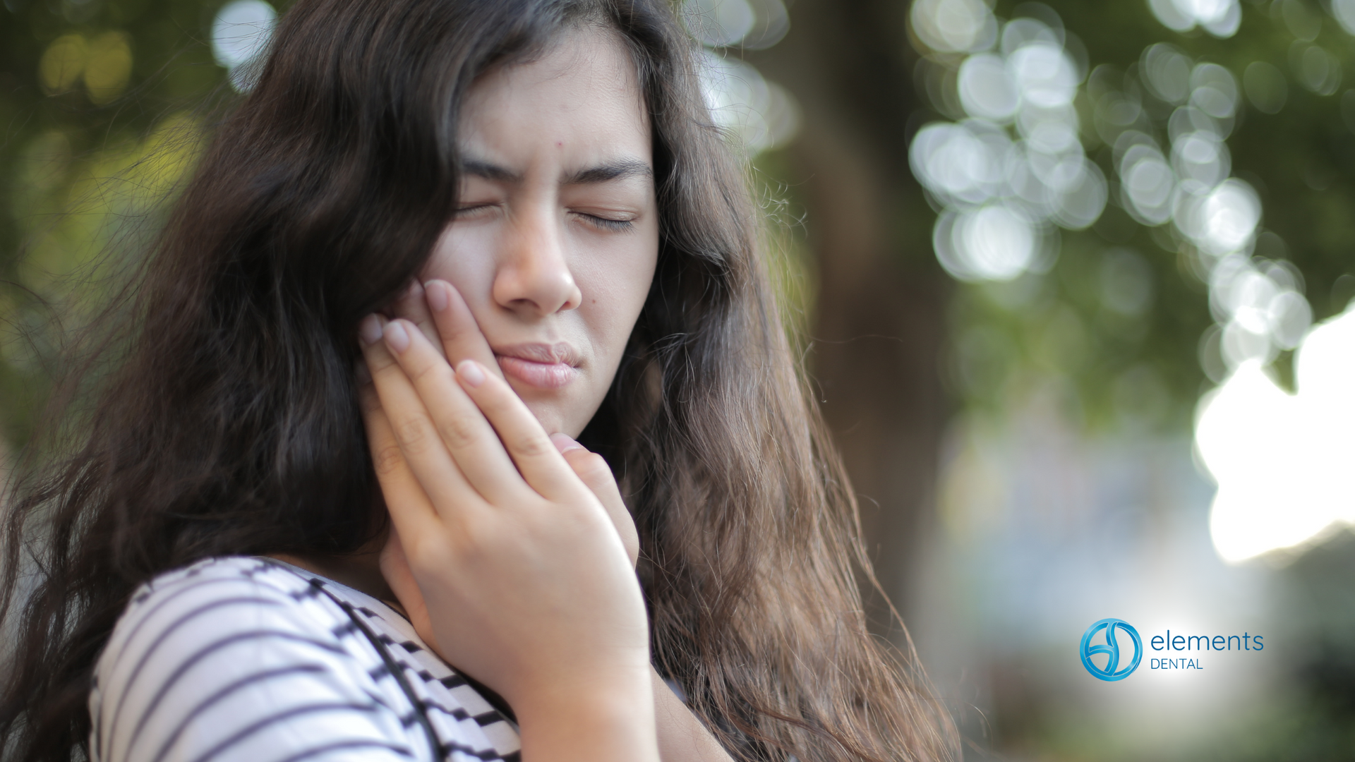 Woman with long hair holding her jaw, eyes closed, showing signs of pain. Outdoors, blurred background.