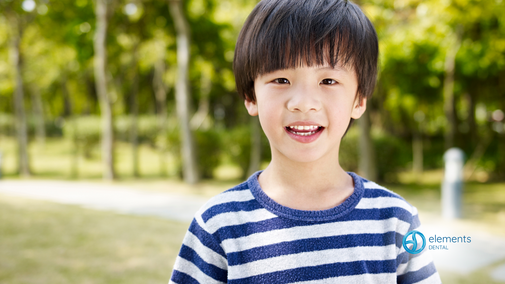 A young boy in a striped shirt is smiling in a park.