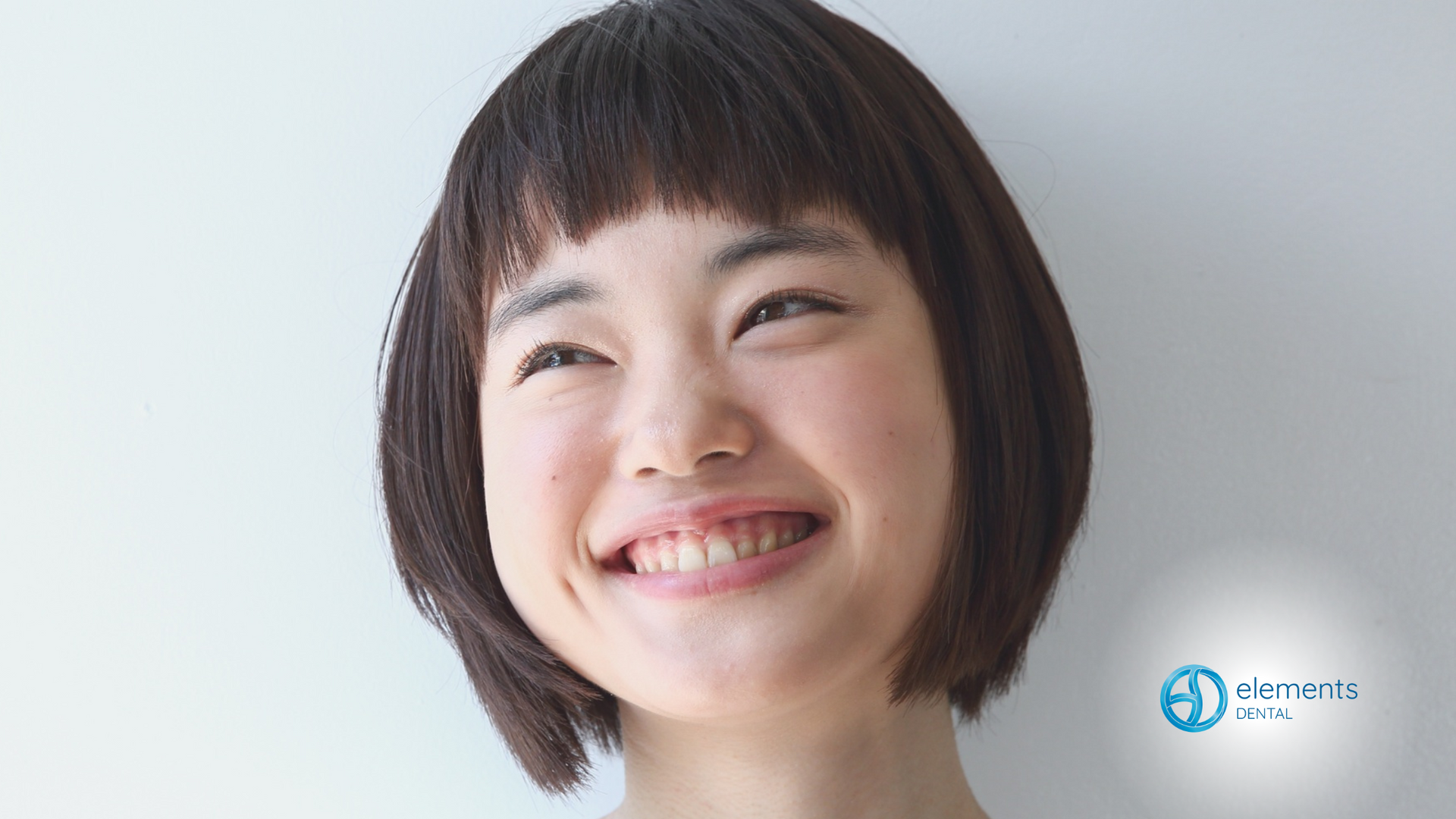 Woman with short, dark hair smiles broadly, leaning slightly toward the camera against a white background.