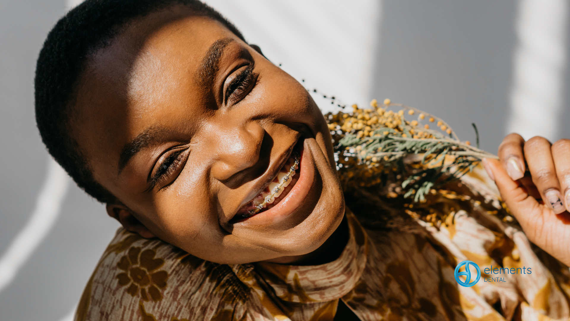 A woman with braces on her teeth is smiling and holding a flower.