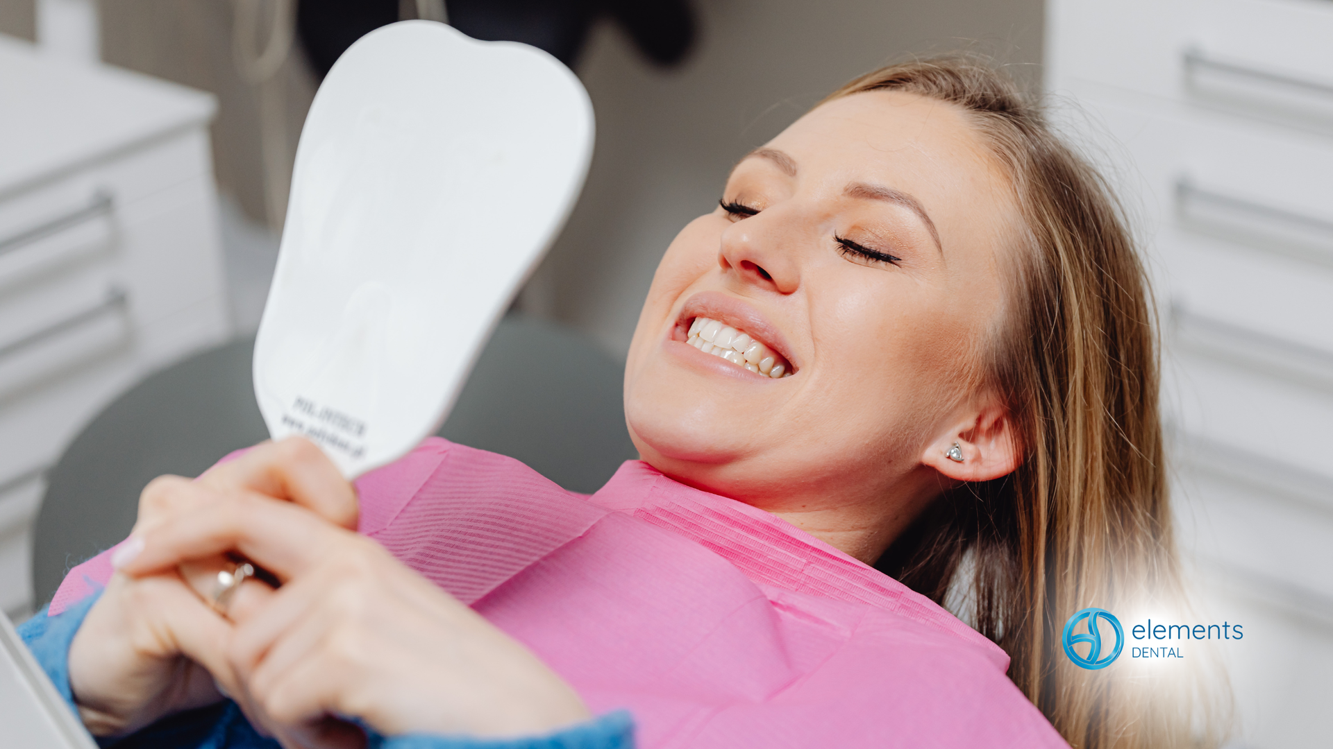 Woman smiling, looking at teeth in a hand mirror in a dental office. She wears a pink bib.