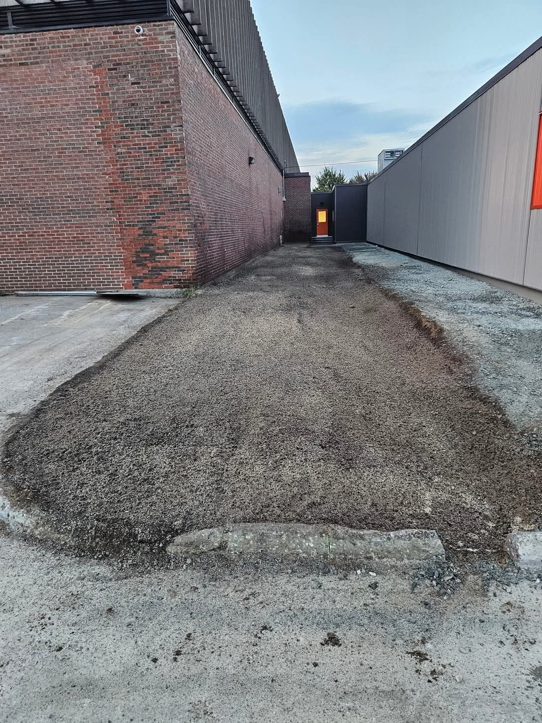 Allée de gravier entre des bâtiments en briques et gris, menant à une porte éclairée.
