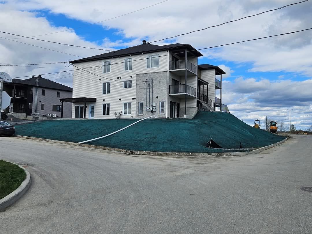 Immeuble d'appartements de trois étages au sommet d'une colline herbeuse, à un carrefour routier, ciel nuageux.
