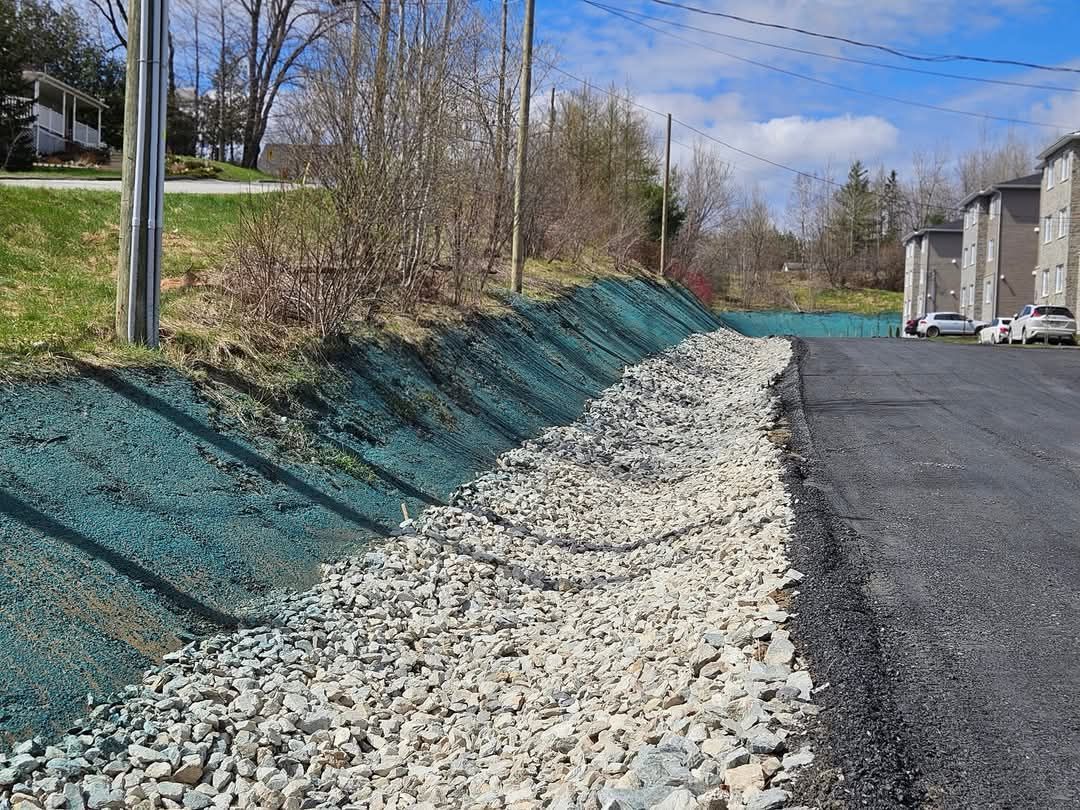 Un fossé de drainage tapissé de gravier longe une route goudronnée. La berge en pente est recouverte d'hydroensemencement bleu.