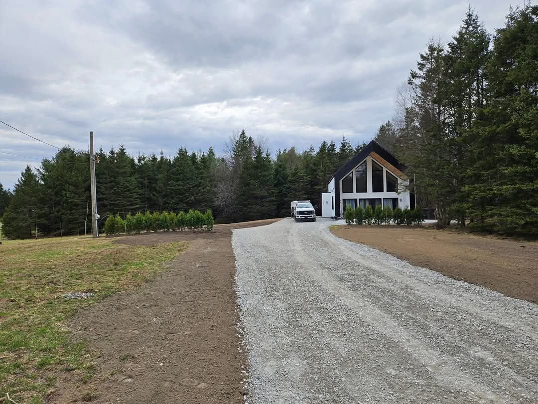 Une allée de gravier mène à une maison blanche moderne aux grandes fenêtres, nichée au milieu des arbres sous un ciel nuageux.