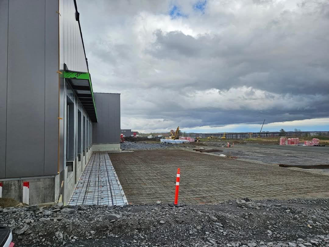 Chantier de construction avec un bâtiment métallique gris, un sol en terre battue et un ciel nuageux.